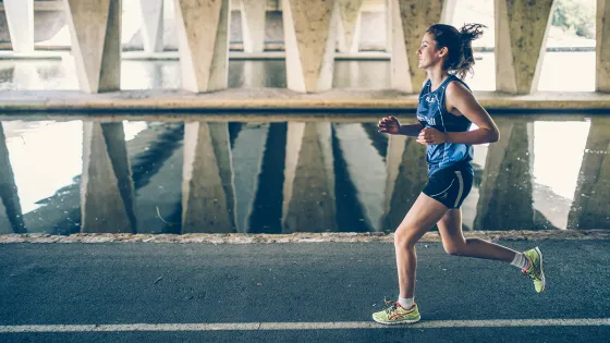 A young woman jogs along a waterfront pathway beneath a concrete bridge, reflecting on the water's surface beside her.