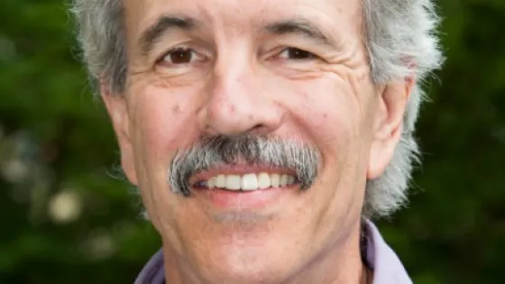 Portrait of a smiling older man with a mustache, wearing a purple shirt, set against a green blurred background.