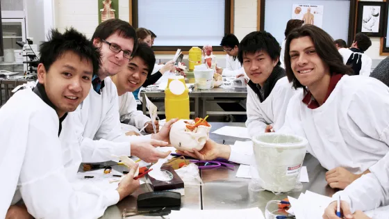 A group of six students in lab coats working together at a laboratory table, with anatomical models and instruments visible.