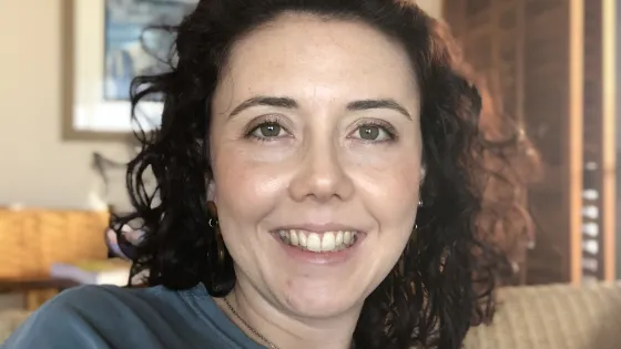A smiling woman with curly dark hair wearing a blue t-shirt, seated indoors.