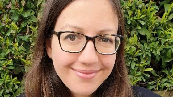 A woman with glasses smiling in front of a lush green hedge.
