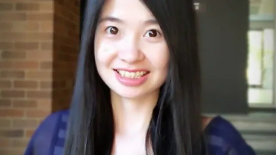 A smiling young woman with long black hair, wearing a navy blue striped top, standing indoors.