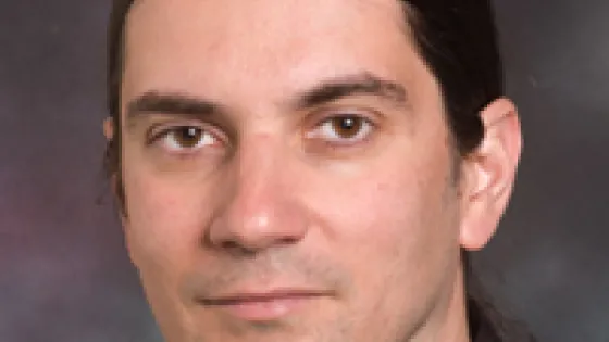 Portrait of a man with long dark hair and a plain expression, wearing a dark shirt with a striped collar.