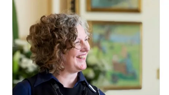 A woman with curly hair smiling and looking to the side in a room with a painting in the background.