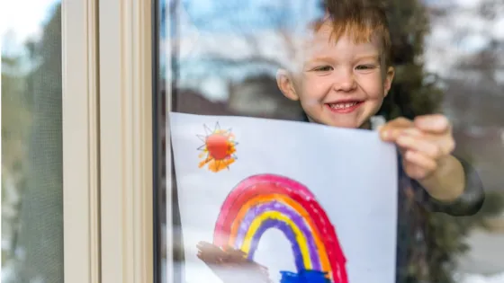 A young boy smiles as he shows off his colorful drawing of a rainbow and sun through a window.