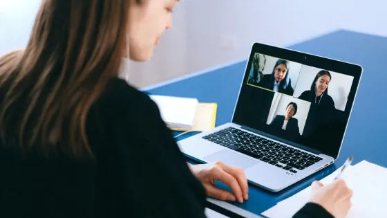 A woman participates in a video call with three other participants displayed on her laptop screen in an office setting.