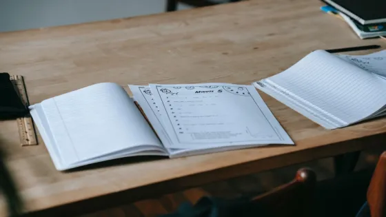 A wooden table holds an open notebook, worksheets, a ruler, and a book, suggesting a study or work setting.