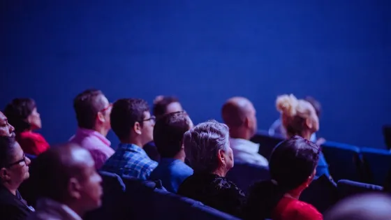 Audience members seated in a theater watching a presentation, focused on the stage ahead.