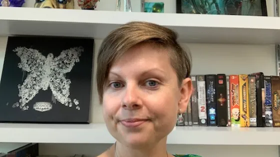 A woman with short hair smiling in front of a bookshelf filled with DVDs and various decorative items.