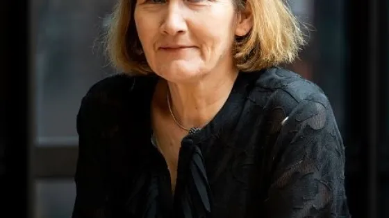 A woman with shoulder-length blonde hair, wearing a black blouse, sits at a table with books, smiling slightly at the camera.