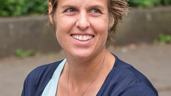 A smiling woman with short blonde hair wearing a blue cardigan and a turquoise top, outdoors.