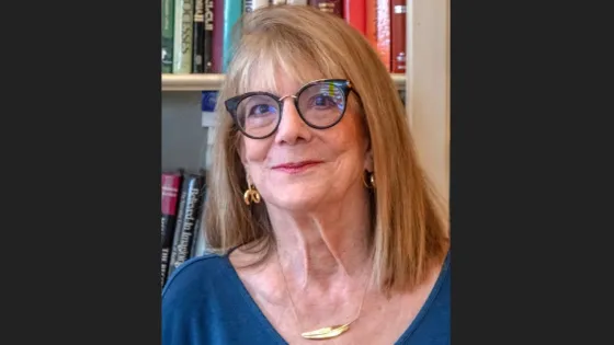 A senior woman with blonde hair and glasses, smiling in front of a bookshelf.