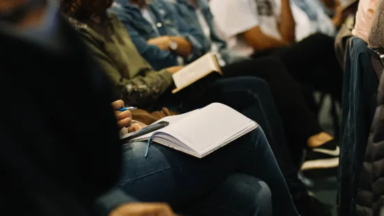 People seated in a lecture hall with notebooks on their laps, focused on writing notes.