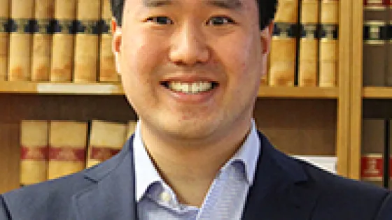 A man in a blue suit and tie smiles in front of bookshelves filled with law books.