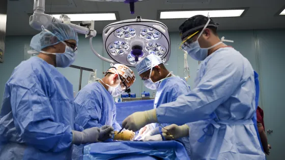 Surgeons in scrubs and surgical masks operate on a patient in a well-lit operating room.