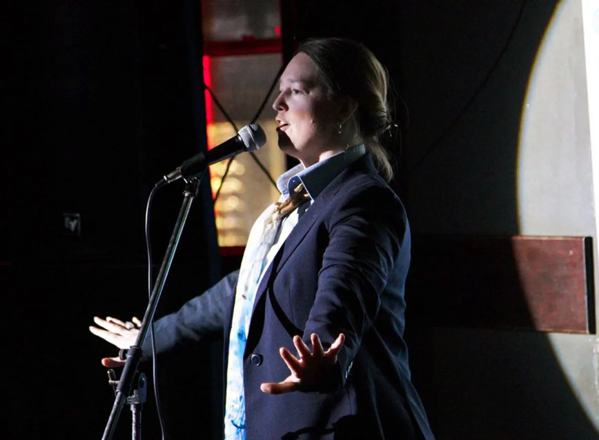 A woman speaking into a microphone on stage, illuminated by dramatic lighting.