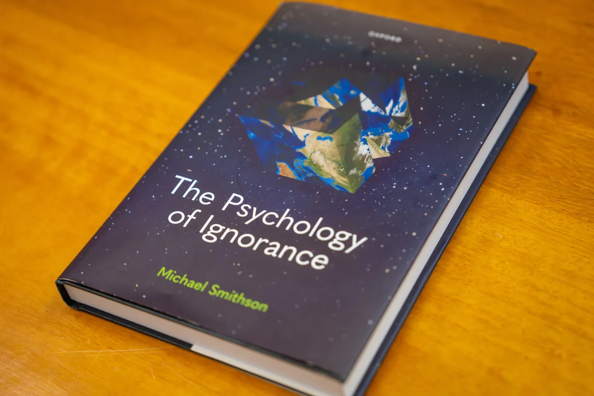 A book titled The Psychology of Ignorance by Micahel Smithson is on a table. The book cover includes a 2D map of the world that is scrunched up to look like a globe