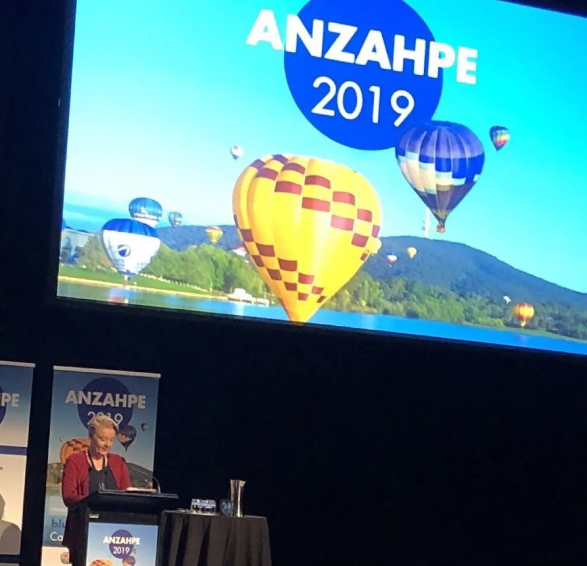 A person standing at a podium in a conference hall, with a large screen behind them displaying an image of colorful hot air balloons and the text "ANZAHPE 2019."