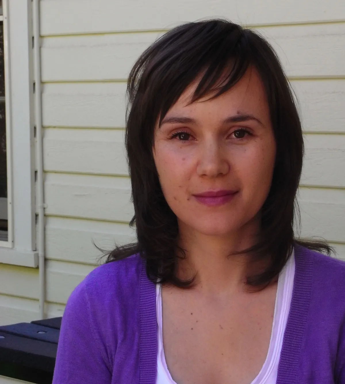 A woman with shoulder-length brown hair and a purple cardigan stands against a yellow house background.