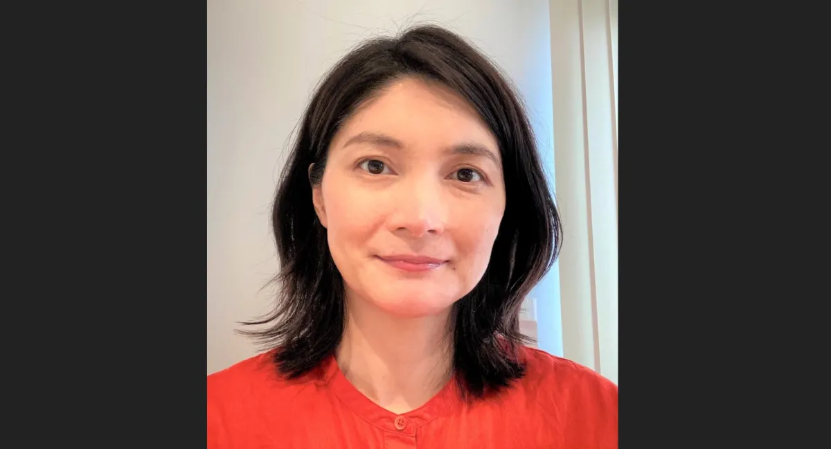 A middle-aged Asian woman with shoulder-length black hair, wearing a red blouse, smiling gently at the camera in an indoor setting.