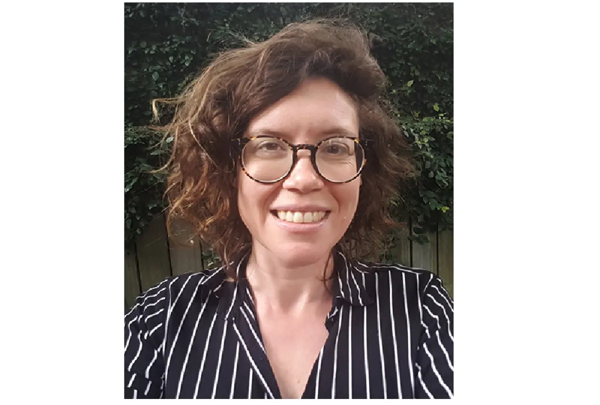 A woman with curly hair and glasses smiling in front of a greenery background, wearing a striped shirt.