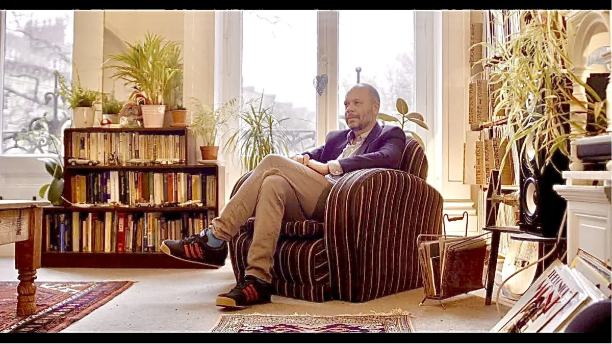 A man sits relaxed in an armchair in a cozy, book-filled room with large windows and various plants.