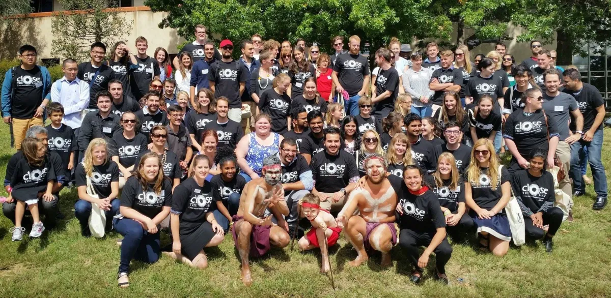 A large group of diverse people wearing matching t-shirts with the text "CLOSE THE GAP" posing together for a group photo outdoors.