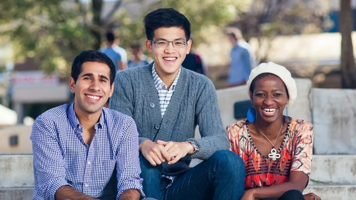 Three young adults, smiling and sitting together outdoors on a sunny day.