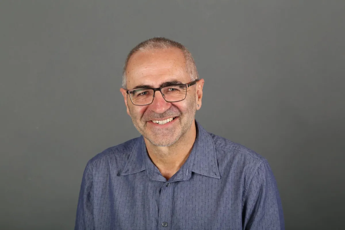 A smiling elderly man with glasses wearing a blue shirt against a gray background.