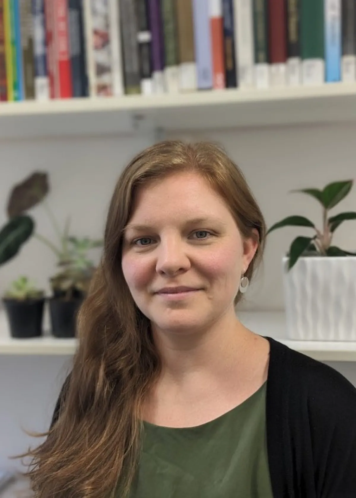 A woman with long brown hair smiling gently, wearing a green top, and standing in front of a bookshelf with plants.