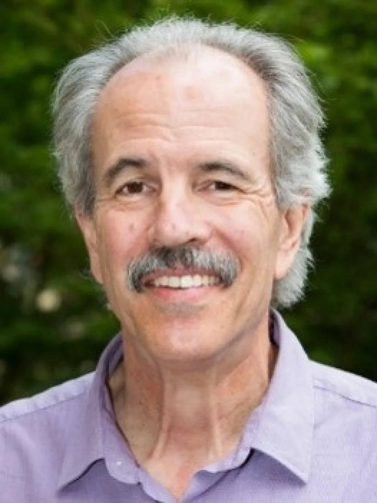 A portrait of an elderly man with a warm smile, sporting a neat gray mustache and wearing a purple shirt, with a blurred green background.
