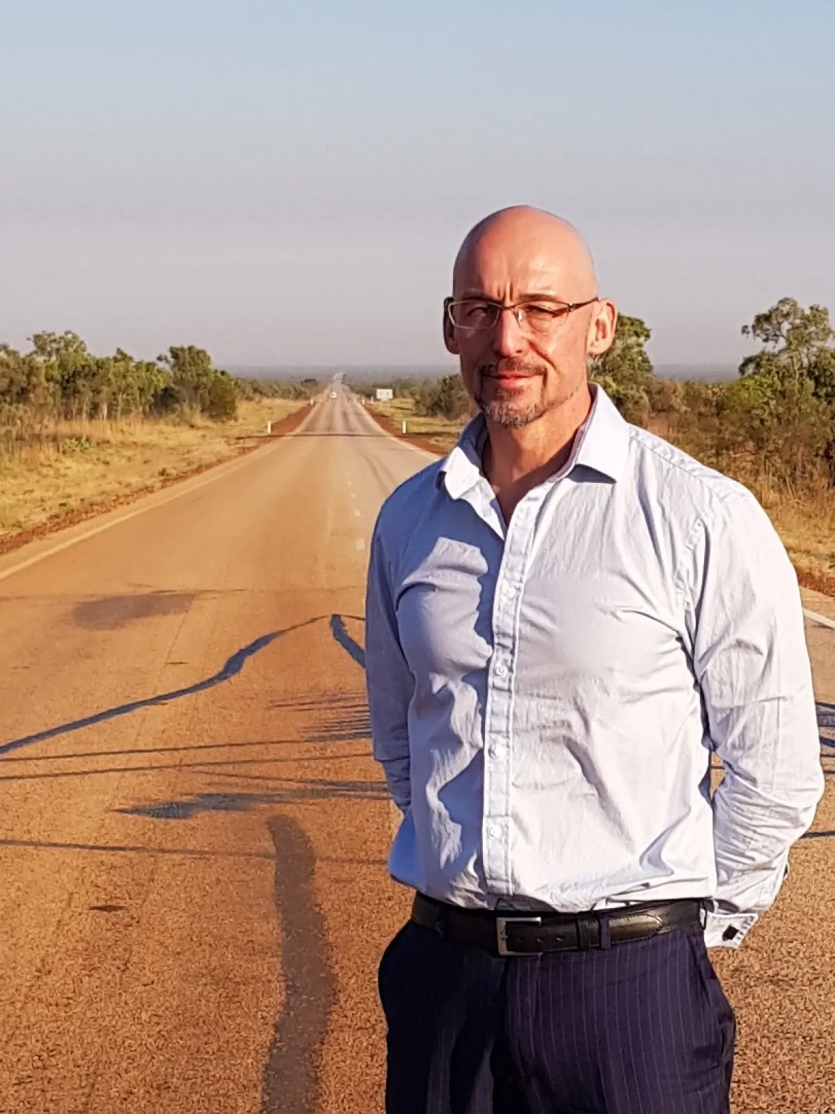 A man stands in the middle of a long, straight road stretching into the distance, wearing glasses and a semi-formal shirt.
