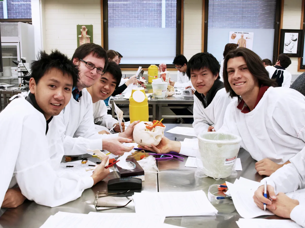 A group of six students in lab coats working together at a laboratory table, with anatomical models and instruments visible.