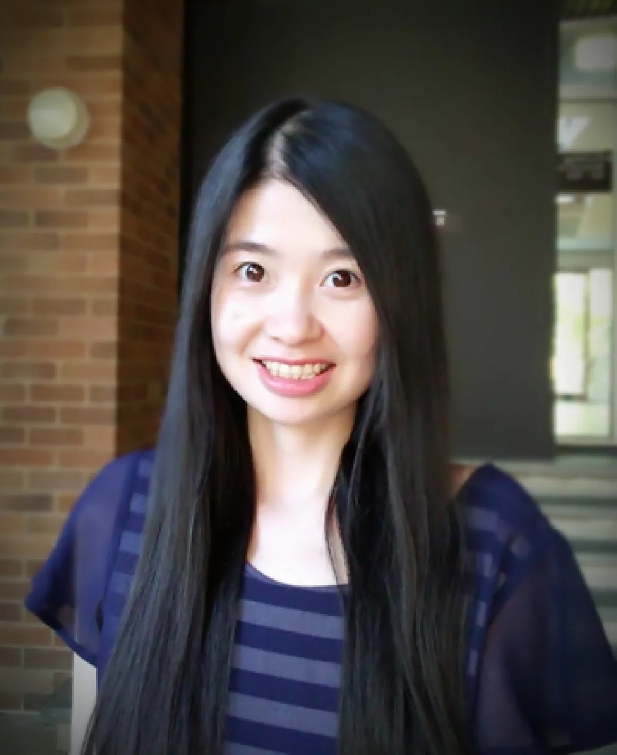 A smiling young woman with long black hair, wearing a navy blue striped top, standing indoors.